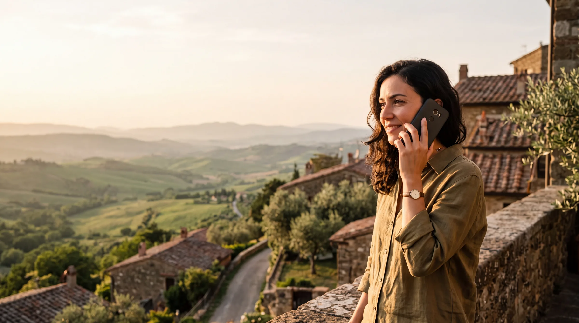 A woman on a stone terrace above a green valley at golden hour, talking on her phone
