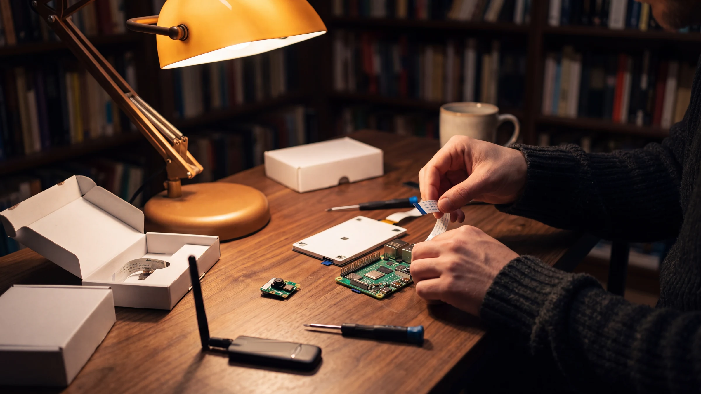 Hands at a lamp-lit wooden workbench connecting a ribbon cable to the kit's board, sealed boxes and the 4G stick beside them