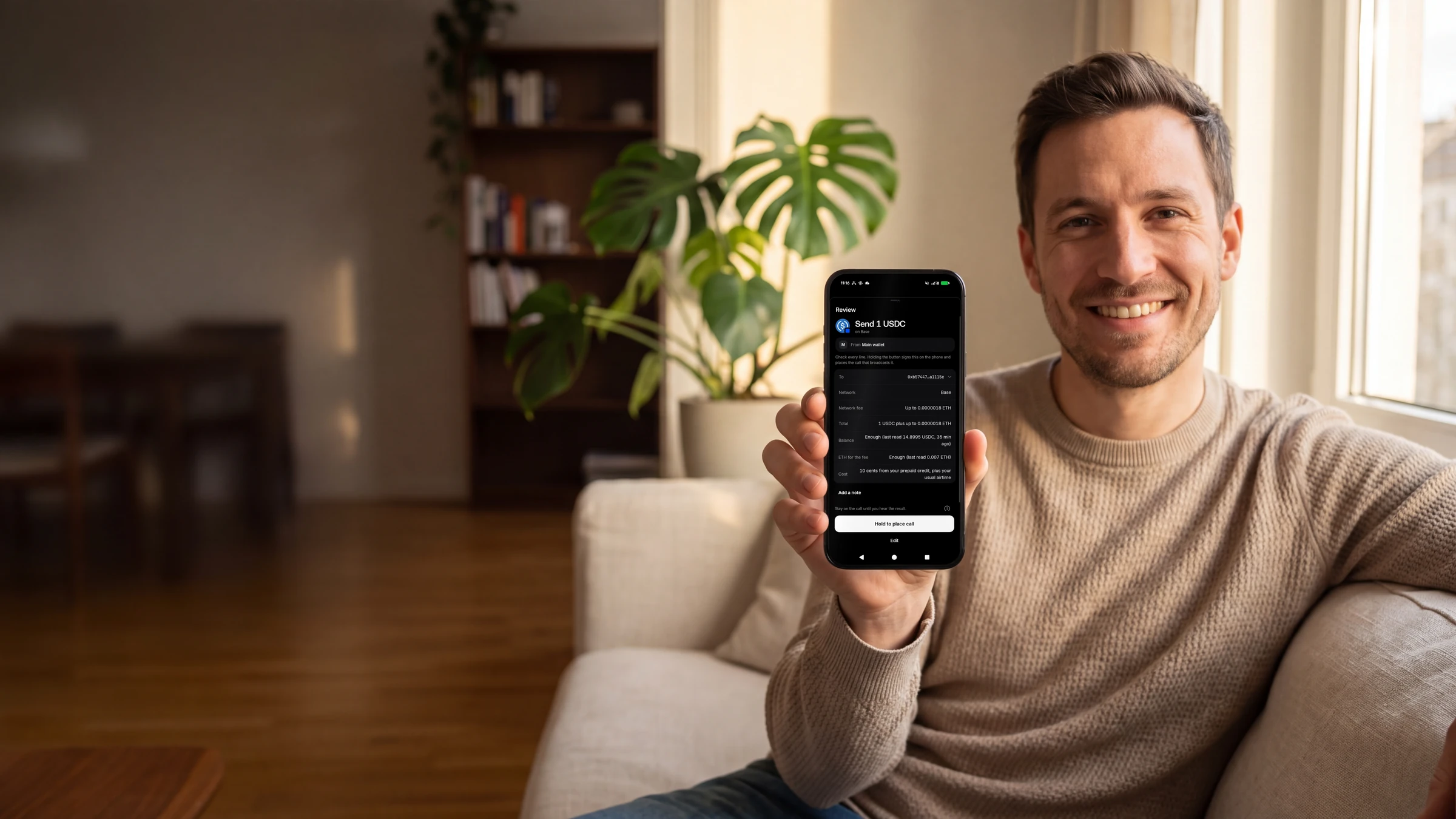 A man on a sofa by a sunlit window holds up his phone, showing the TELRPC Wallet review sheet for sending 1 USDC on Base