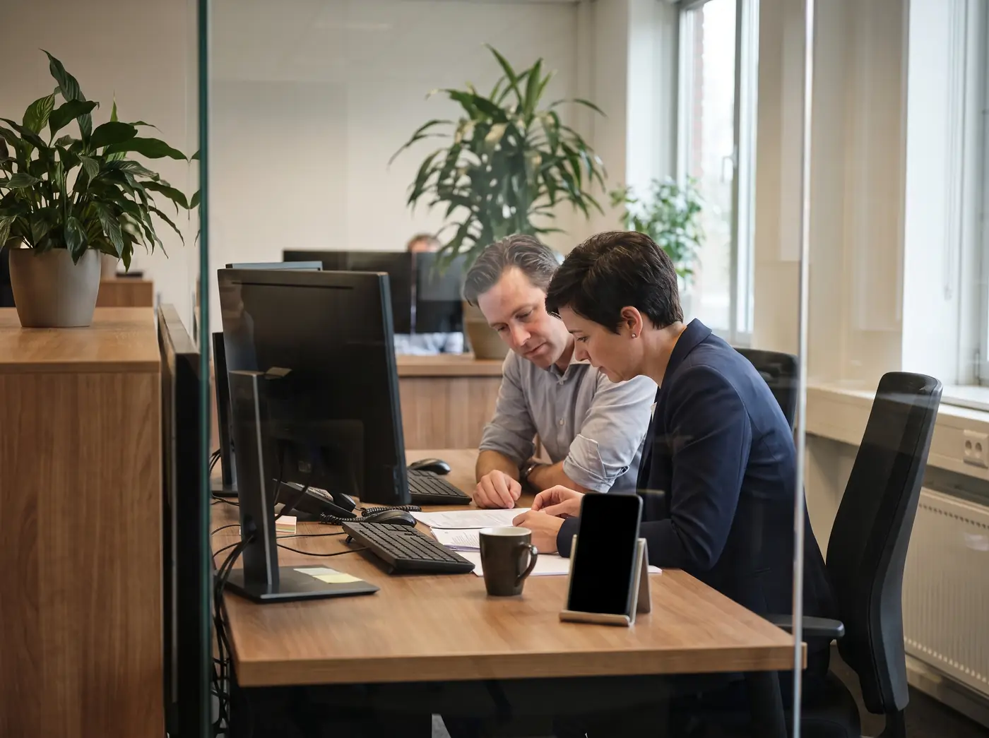 Two treasury professionals at a desk in a daylit office reviewing a document together, a dedicated handset standing beside them with its screen dark