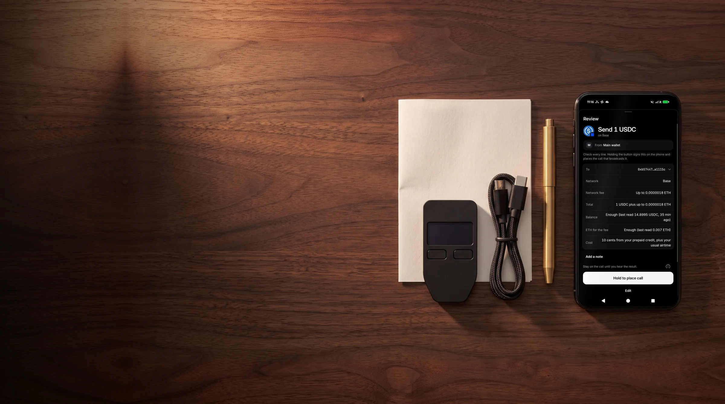 A hardware wallet on a walnut desk beside a phone showing the TELRPC Wallet review sheet: send 1 USDC on Base, hold to place the call